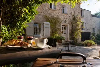 Outdoor breakfast table with croissants and coffee in a tranquil sunlit garden, guesthouse in background, suitcase nearby