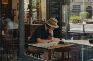 Traveler journaling alone at a table in a cozy Marne-la-Vallée café, afternoon sunlight streaming through the window, with a subtle view of the quiet street outside.