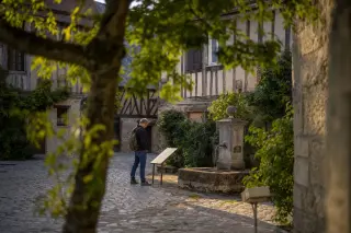 Man quietly reading a plaque by an old stone fountain, surrounded by historic houses in gentle evening light, viewed through tree branches