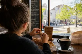 Person sitting alone in a sunlit bakery, breaking bread at a rustic table, with local buildings and greenery visible through the window.